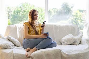 Young woman lying on the couch at home using smartphone - AFVF06268