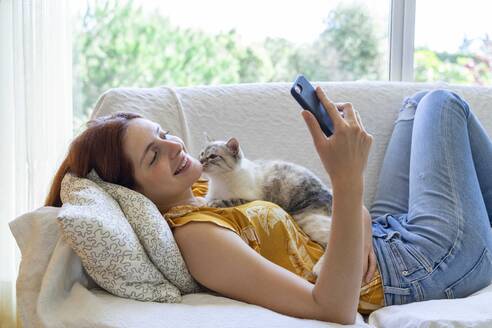 Glückliche junge Frau, die mit ihrer Katze zu Hause auf der Couch liegt und ihr Smartphone benutzt - AFVF06273