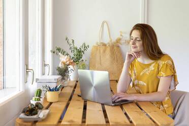 Young woman sitting at table at home using laptop - AFVF06284