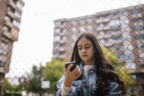 Niedriger Winkel Blick auf Teenager-Mädchen mit Smartphone, während stehend gegen Maschendrahtzaun - GRCF00220