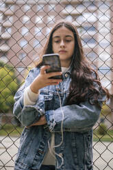 Selbstbewusstes Teenager-Mädchen, das sein Smartphone am Maschendrahtzaun des Sportplatzes benutzt - GRCF00221