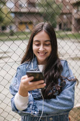 Smiling teenage girl using mobile phone while standing against chainlink fence at sports court - GRCF00222