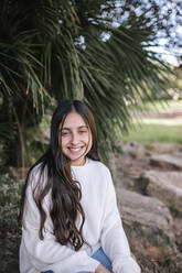 Portrait of smiling teenage girl sitting against palm tree at park - GRCF00226