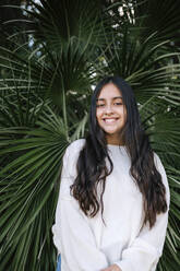 Portrait of happy teenage girl with long brown hair standing against palm tree at park - GRCF00228