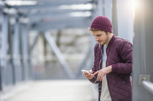 Junger Mann auf einer Brücke mit Ohrstöpseln und Smartphone - DIGF10901