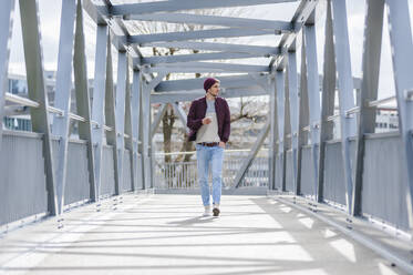 Junger Mann, der mit Ohrstöpseln und Smartphone auf einer Brücke spazieren geht - DIGF10902