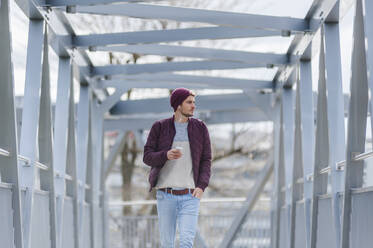 Junger Mann, der mit Ohrstöpseln und Smartphone auf einer Brücke spazieren geht - DIGF10903