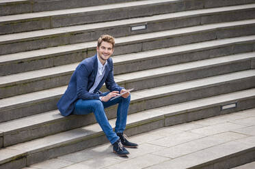 Glücklicher junger Geschäftsmann sitzt auf einer Treppe und benutzt ein Tablet - DIGF10929