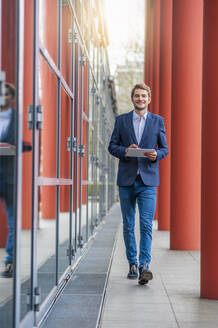 Junger Geschäftsmann mit Tablet zu Fuß außerhalb eines Gebäudes - DIGF10935