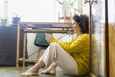 Smiling woman sitting on the floor at home wearing headphones and using smartphone - ERRF03802