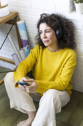 Frau sitzt zu Hause auf dem Boden, trägt Kopfhörer und benutzt ein Smartphone - ERRF03803