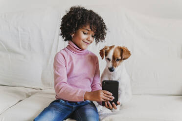 Portrait of smiling little girl sitting on the couch with her dog taking selfie with smartphone - EBBF00009