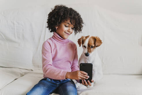 Porträt eines lächelnden kleinen Mädchens, das mit ihrem Hund auf der Couch sitzt und ein Selfie mit dem Smartphone macht - EBBF00009