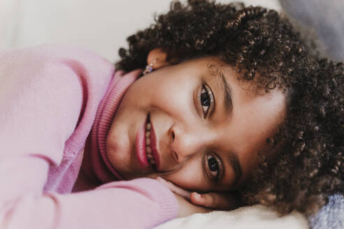 Portrait of smiling little girl wearing pink turtleneck pullover lying on couch - EBBF00017