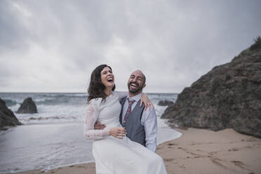 Lachendes Brautpaar am Strand, das seine Frau im Arm hält - SNF00220
