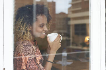 Lächelnde junge Frau mit einer Tasse Kaffee hinter einer Fensterscheibe - VEGF02257