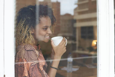 Smiling young woman with cup of coffee behind windowpane - VEGF02257