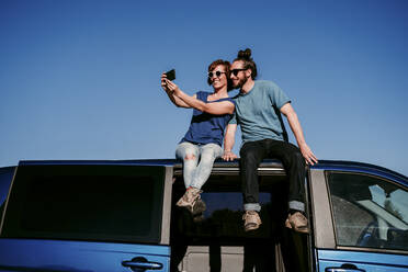 Glückliches Paar, das auf dem Dach eines Minivans sitzt und ein Selfie macht - EBBF00059