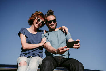 Happy couple sitting on the roof of a minivan taking a selfie - EBBF00060