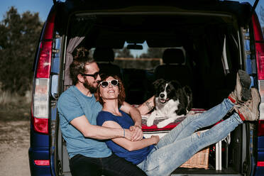 Happy couple with dog sitting in the boot of a minivan - EBBF00065