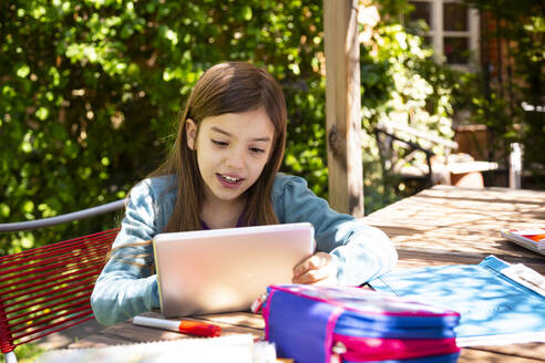 Girl sitting at garden table doing homework and using tablet - LVF08894