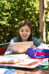 Mädchen sitzt am Gartentisch, macht Hausaufgaben und benutzt ein Tablet - LVF08895