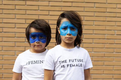 Porträt von zwei Brüdern mit aufgemalten blauen Masken im Gesicht, die T-Shirts mit feministischen Aufdrucken tragen, lizenzfreies Stockfoto