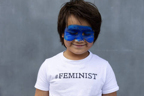 Porträt eines lächelnden Jungen mit gemalter blauer Maske im Gesicht, der ein T-Shirt mit dem Aufdruck 'Feminist' trägt, lizenzfreies Stockfoto