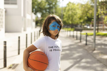 Porträt eines Jungen mit gemalter blauer Maske im Gesicht, der einen Basketball hält - VABF02937