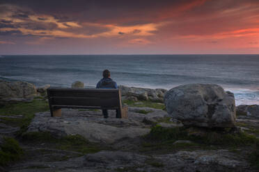 Mann beobachtet einen atemberaubenden Sonnenuntergang auf einer Bank an der Küste sitzend - CAVF80924