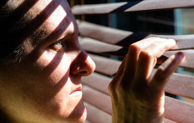 Erwachsene Frau mit blauen Augen, die mit ihren Fingern einen hölzernen Vorhang öffnet, um durch das Fenster zu schauen, während das Sonnenlicht Sonne und Schatten auf ihrem Gesicht erzeugt. Horizontales Foto. - CAVF80966