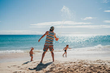 Kinder, die mit ihrem Vater an einem sonnigen Tag am Strand mit Wasser spielen - CAVF80988