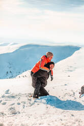 Ein Mann im Skianzug kniet im Schnee und nimmt eine Frau huckepack, lacht und schaut in die Kamera. - CUF55098