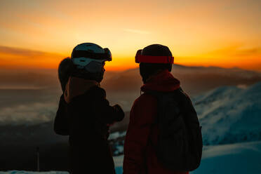 Silhouette von zwei Personen mit Skihelmen und -brillen bei Sonnenuntergang auf einem Berg im Winter - CUF55105