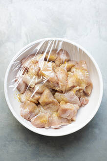 Weiße Schüssel mit gewürfeltem rohem Hühnerfleisch, das mit Gewürzen bestreut und mit Frischhaltefolie abgedeckt ist. - CUF55106