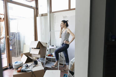 Woman standing with one foot on the bottom of a stepladder talking on a phone with cardboard boxes nearby. - CUF55136