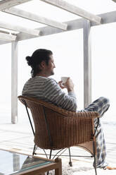 A man sitting in a rattan chair on a deck looking into the distance. - CUF55138