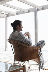 Ein Mann sitzt in einem Rattansessel auf einer Terrasse und schaut in die Ferne. - CUF55138