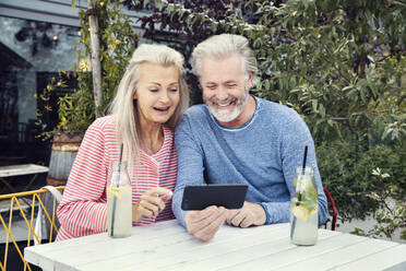 Ein Paar sitzt an einem Tisch in einem Garten mit Getränken in Flaschen und schaut gemeinsam auf ein Handy. - CUF55180