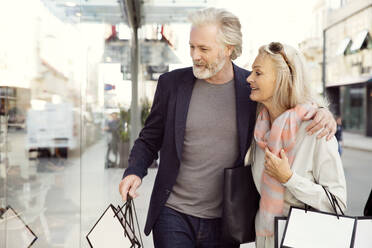 A couple with arms around each other carrying shopping bags and looking in a shop window. - CUF55192