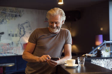 Ein Mann lehnt an einer Theke neben einer Kaffeetasse, lächelt und schaut auf ein Mobiltelefon. - CUF55197