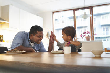 Ein Vater und sein Sohn sitzen am Küchentisch, lächeln und geben sich gegenseitig ein High Five. - CUF55231