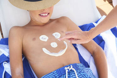 A boy lying on a sun lounger with a woman drawing a face on his chest with sun cream. - CUF55246