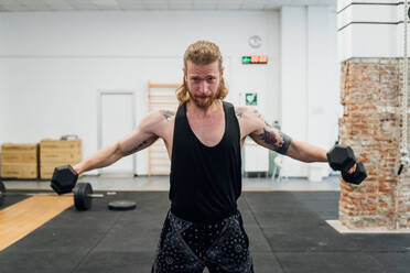 Man standing in a gym with tattooed arms looking at the camera holding small dumbells with outstretched arms. - CUF55265