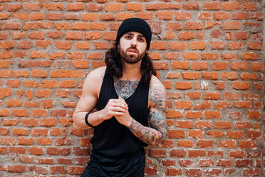 A man wearing a vest and woolly hat with tattooed arms and chest leaning against a brick wall. - CUF55278