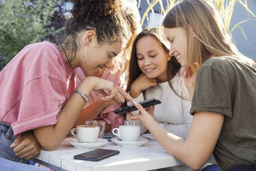 Drei Frauen sitzen an einem Tisch, schauen auf ein Handy und lächeln. - CUF55290