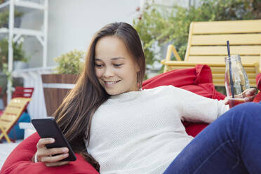 A woman lounging on a beanbag with a bottle of drink, looking at a mobile phone. - CUF55294