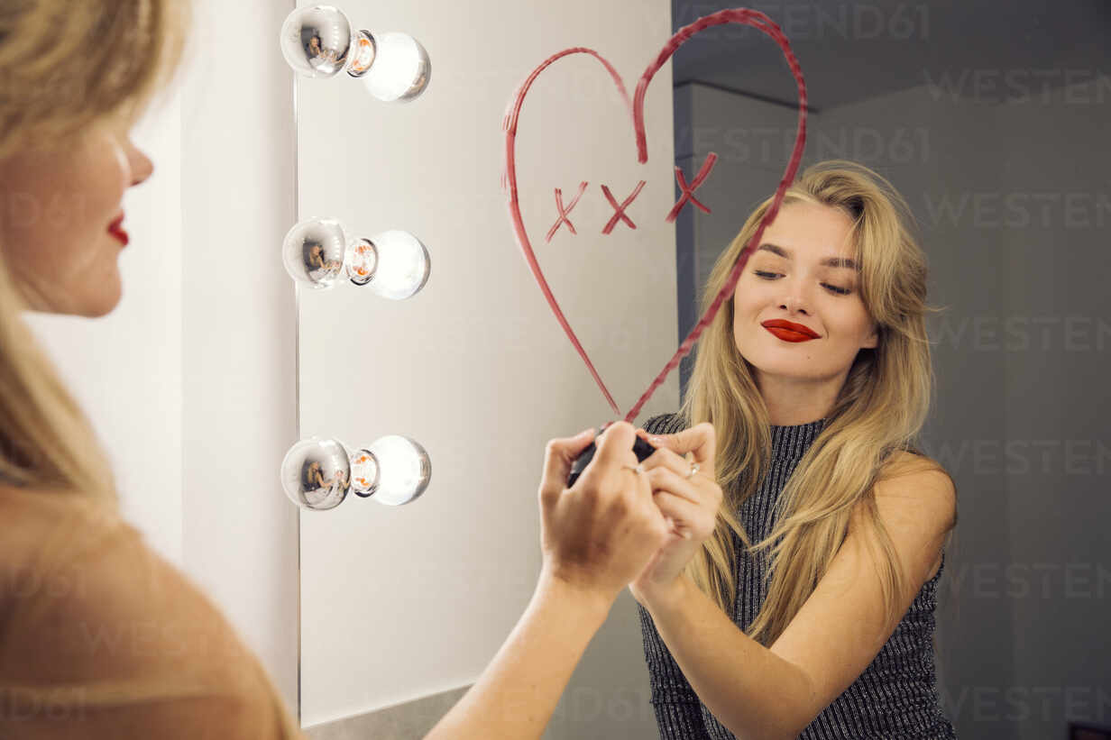 Eine Frau schaut in einen Badezimmerspiegel und malt mit Lippenstift ein  Herz auf den Spiegel., lizenzfreies Stockfoto
