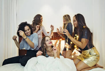 Fünf Frauen, die zum Ausgehen gekleidet sind, sitzen und liegen auf einem Bett, eine hält eine Lichterkette. - CUF55306