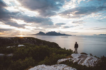 Mann steht auf einem Felsen bei Sonnenuntergang am Meer und schaut auf eine Insel - CAVF81072